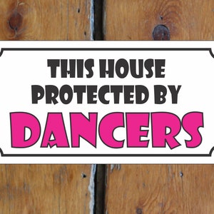 May include: White sign with black border and rounded corners. The sign reads "This house protected by dancers" in black text. The word "dancers" is in pink text.