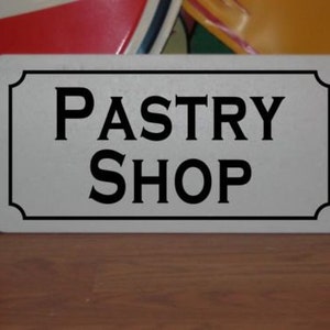 May include: White metal sign with black lettering that reads "Pastry Shop".