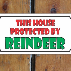Puede incluir: Señal blanca con borde negro y esquinas redondeadas. La señal dice "Esta casa protegida por renos" en letras rojas y verdes.