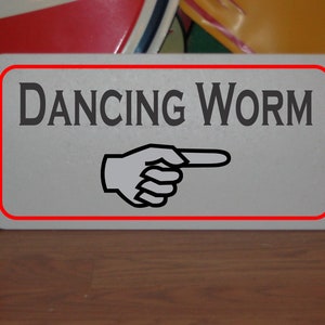 May include: A white sign with a red border and black text that reads "DANCING WORM". A black pointing hand is below the text.