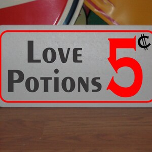 以下が含まれることがあります： 赤い縁取りと赤い文字で「Love Potions 5¢」と書かれた白い看板です。