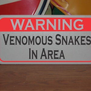 Peut inclure: Un panneau d'avertissement blanc avec une bordure rouge. Le panneau indique "ATTENTION SERPENTS VENIMEUX DANS LA ZONE".