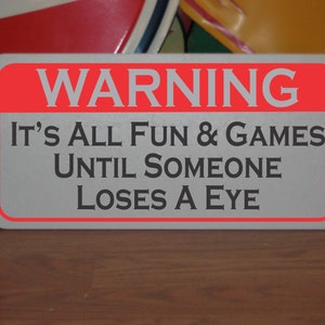 May include: A white sign with a red border and the text "WARNING IT'S ALL FUN & GAMES UNTIL SOMEONE LOSES AN EYE" in black.