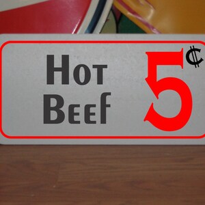 May include: A white sign with red trim and black text that reads "Hot Beef 5¢". The sign is on a wooden surface.