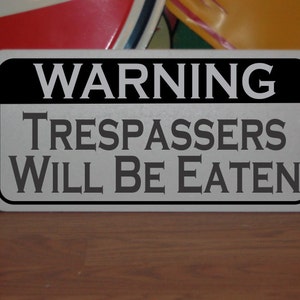 Puede incluir: Un cartel de advertencia en blanco y negro con el texto "WARNING TRESPASSERS WILL BE EATEN".