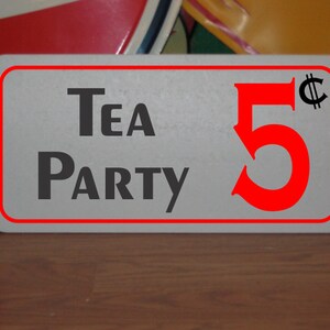 Könnte beinhalten: Ein weißes rechteckiges Schild mit rotem Rand und schwarzem Text, auf dem "Tea Party 5¢" steht. Die Zahl 5 ist rot und das Cent-Symbol ist schwarz.
