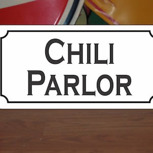 May include: White sign with black border and ornate corners. The sign reads "CHILI PARLOR" in black serif font.