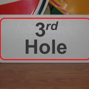 以下が含まれることがあります： ゴルフコースの3番ホールを示す、赤い縁取りのある白い看板です。