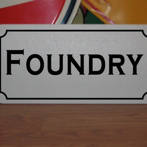 May include: White rectangular sign with black border and rounded corners. The sign says "FOUNDRY" in black letters.