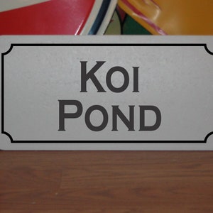 Op de afbeelding: Een rechthoekig bord met de woorden "Koi Pond" in donkergrijze letters. Het bord heeft een lichtgrijze achtergrond met een zwarte rand. Het bord staat op een houten oppervlak.
