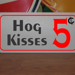 Puede incluir: Un letrero blanco con borde rojo y texto negro que dice "Hog Kisses 5¢". El número 5 está en una fuente roja grande.