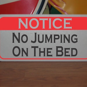 May include: A white sign with a red border and the text "NOTICE NO JUMPING ON THE BED" in black.