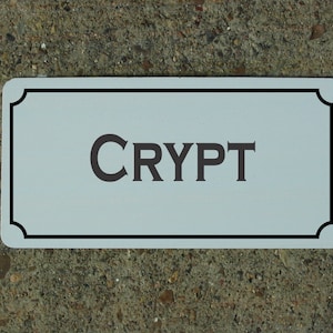 May include: A white rectangular sign with a black border and rounded corners. The sign has the word "CRYPT" printed in black letters.
