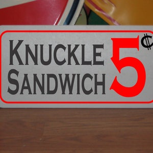 May include: A white sign with red lettering advertising a "Knuckle Sandwich" for 5 cents. The sign has a red border and is slightly worn.