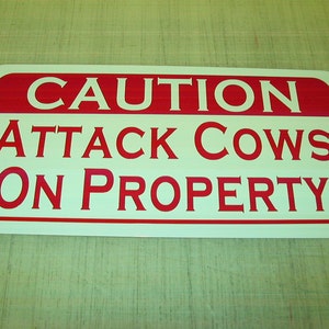 Könnte beinhalten: Ein weißes Schild mit roter Aufschrift, auf dem "ACHTUNG ANGRIFFSKÜHE AUF GRUNDSTÜCK" steht.