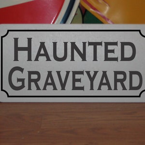 Puede incluir: Un letrero blanco con letras negras que dice "HAUNTED GRAVEYARD". El letrero tiene un borde decorativo y está sobre una superficie de madera.