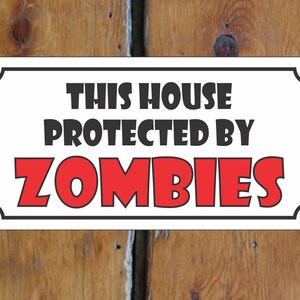 Puede incluir: Un cartel blanco con borde negro y esquinas redondeadas. El cartel dice "This house protected by ZOMBIES" en letras negras y rojas.
