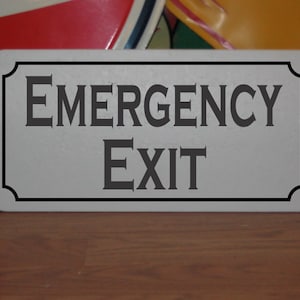 May include: A white rectangular sign with a black border and rounded corners. The sign has black text that reads "EMERGENCY EXIT".
