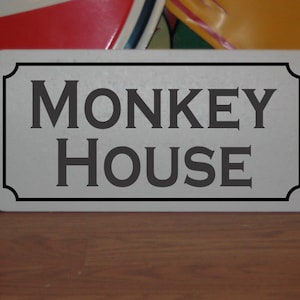 May include: A rectangular sign with the words "MONKEY HOUSE" in dark gray, centered on a light gray background. The sign has a black border and is likely for decorative purposes.