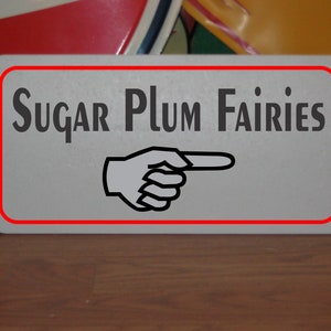 May include: A white sign with a red border and black text that reads "Sugar Plum Fairies". A black and white pointing hand is below the text.