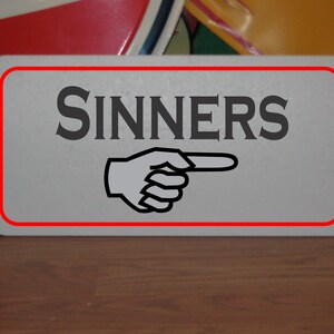 May include: A white sign with a red border that says "SINNERS" in black letters with a black pointing hand pointing to the right.