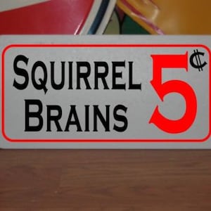 Op de afbeelding: Wit metalen bord met een rode rand en zwarte tekst. Het bord staat "Squirrel Brains 5¢".
