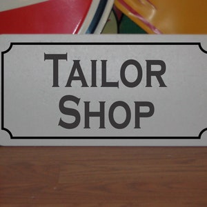 以下が含まれることがあります： 黒い縁取りのある白い金属製の看板に、黒い文字で「TAILOR SHOP」と書かれている。