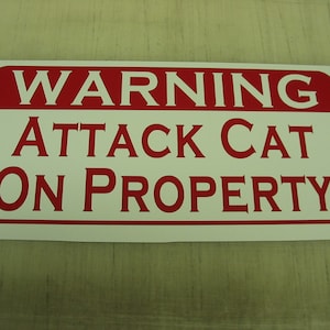 Könnte beinhalten: Ein weißes Schild mit rotem Text, auf dem steht "WARNUNG ANGRIFFSKATZE AUF GRUNDSTÜCK".