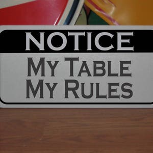 May include: A silver metal sign with black lettering that reads "NOTICE MY TABLE MY RULES".