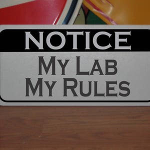May include: A silver metal sign with black lettering that reads "NOTICE MY LAB MY RULES".