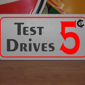 Op de afbeelding: Een wit bord met een rode rand en zwarte tekst die "Test Drives 5¢" zegt. Het getal 5 is in een groot rood lettertype en het centsymbool is zwart.