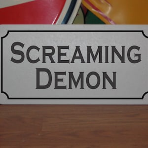 May include: A white rectangular sign with black trim and rounded corners. The sign has black text that reads "SCREAMING DEMON".