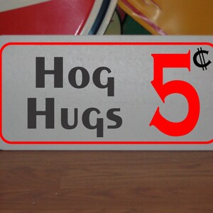 Puede incluir: Un letrero blanco con borde rojo y texto negro que dice "Hog Hugs 5¢". El número 5 es rojo y el símbolo del centavo es negro.