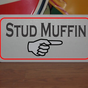 May include: A white sign with a red border and the text "STUD MUFFIN" in black letters. A black and white hand points to the right.