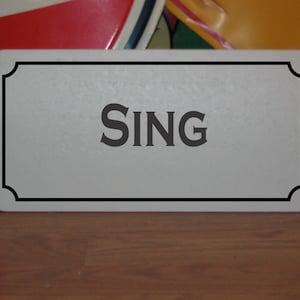 May include: A white sign with a black border and the word "SING" in black lettering.