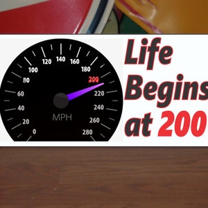 Op de afbeelding: Een wit bord met een zwarte snelheidsmeter waarop "Life Begins at 200" staat. De naald van de snelheidsmeter wijst naar 200 mph.