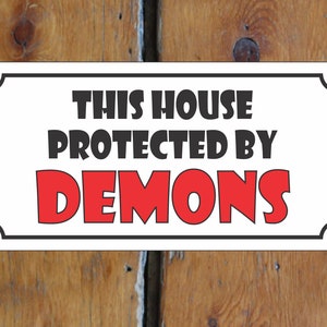 Puede incluir: Un cartel blanco con borde negro y esquinas redondeadas. El cartel dice "This house protected by demons" en letras negras y rojas.