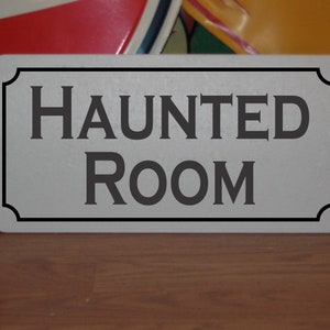 Puede incluir: Un letrero blanco con borde negro y el texto "HAUNTED ROOM" en letras negras.