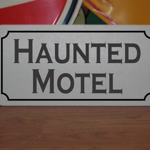 Puede incluir: Un letrero blanco con letras negras que dice "HAUNTED MOTEL". El letrero tiene un borde decorativo.