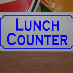 Op de afbeelding: Een wit bord met blauwe tekst met de tekst "Lunch Counter".