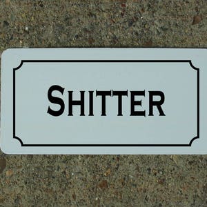 May include: A white rectangular sign with a black border and the word "SHITTER" printed in black letters.