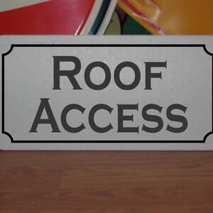 May include: A silver metal sign with black lettering that reads "ROOF ACCESS".