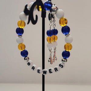 May include: A beaded bracelet and matching dangle earrings set featuring blue, yellow, and white round beads. The bracelet includes white letter beads spelling BREWERS and silver-toned accents. The earrings feature matching beads and silver charms.