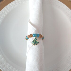 May include: A white cloth napkin rolled and held by a beaded napkin ring. The ring features blue and brown beads and a green fish charm. The napkin rests on a white plate with a gold rim, creating an elegant table setting.
