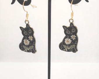 May include: Black cat earrings with daisy accents. These dangle earrings feature a black cat design with white daisy flowers and green leaves. The earrings are suspended from gold-colored hooks and are displayed on a black metal stand.
