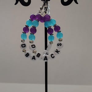 May include: A pair of beaded earrings with a unique design. The earrings feature turquoise and purple beads, with white letter beads spelling out "DO BACKS". The earrings are displayed on a black metal stand.