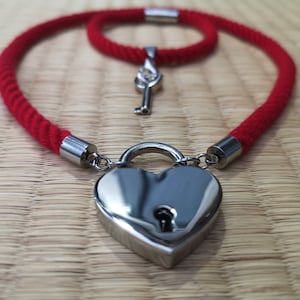 Op de afbeelding: Een rode touw choker ketting met een hartvormig hangslot en een sleutelbedel. Het hangslot is zilverkleurig en heeft een sleutelgat. De ketting ligt op een houten oppervlak.