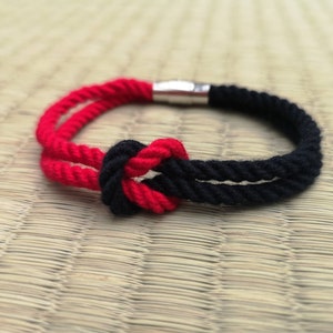 May include: A red and black rope bracelet with a knot design. The bracelet has a silver clasp and is shown on a textured, light brown surface. The rope is thick and appears to be made of a durable material.
