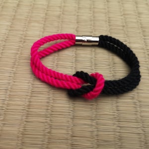 Op de afbeelding: Een roze en zwarte touw armband met een zilveren magnetische sluiting. De armband is in een knoop geknoopt.