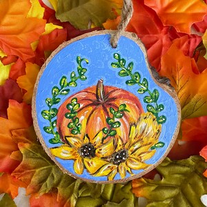 May include: A wooden round ornament painted with a blue background, a pumpkin with orange and yellow hues, and two sunflowers with yellow petals and brown centers. Green leaves surround the pumpkin and sunflowers.